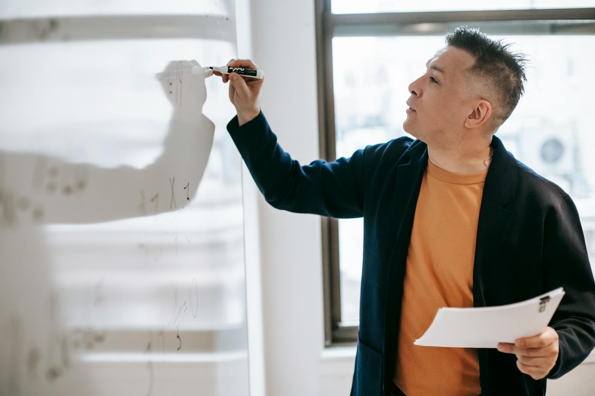 A male educator writing equations on a whiteboard, teaching in a classroom setting.