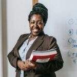 Cheerful African American female teacher wearing formal clothes standing with stacked notebooks in hands near whiteboard in classroom and looking at camera with toothy smile